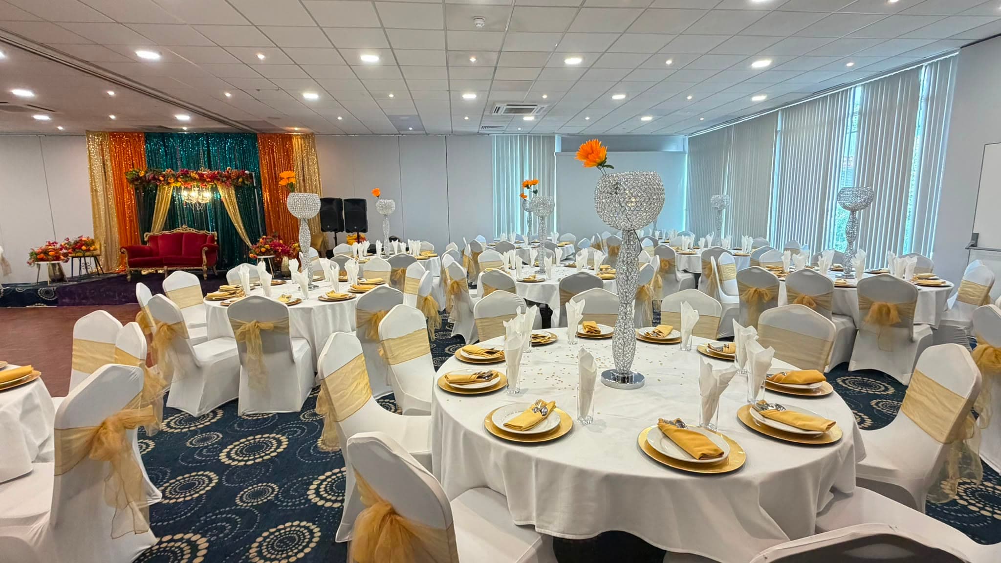 Round tables laid with gold charger plates and crystal centrepieces, with the wedding stage at the far end of the suite.