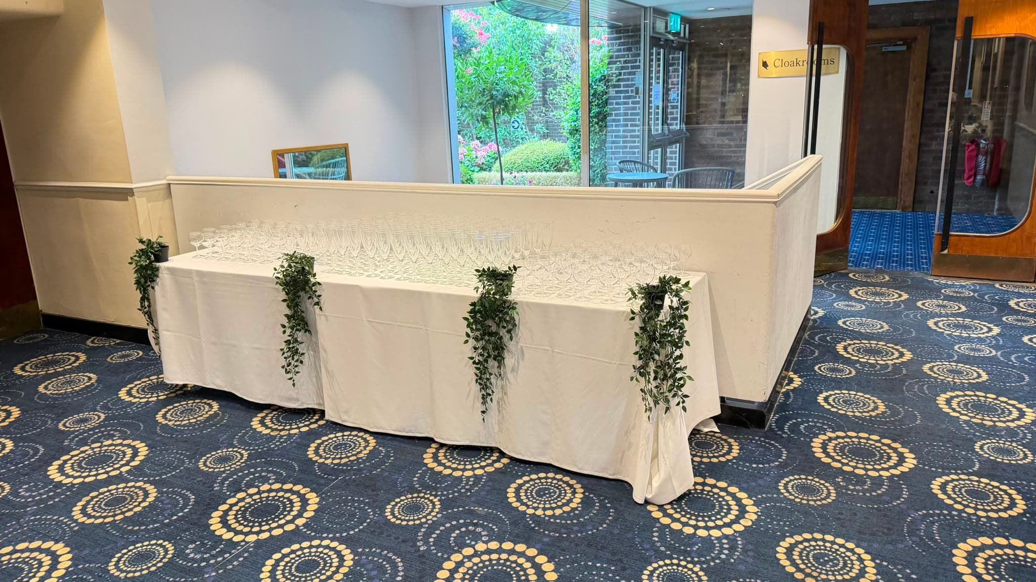 A long dressed table in the foyer lined with glassware for a drinks reception, trailing greenery down the front.