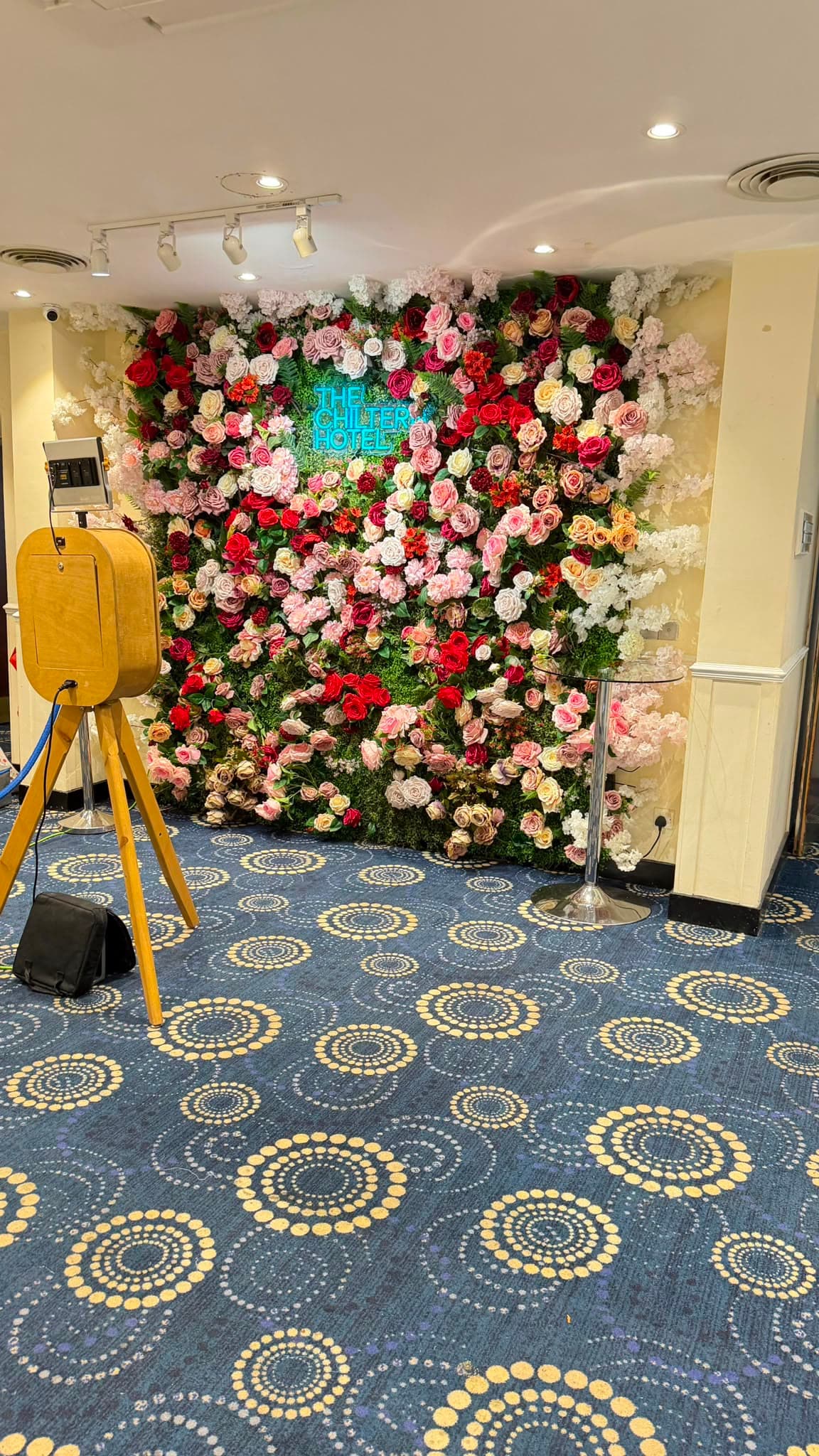 A floor-to-ceiling flower wall of pink, red and cream roses with a neon Chiltern Hotel sign at its centre, and a photo booth set up beside it.