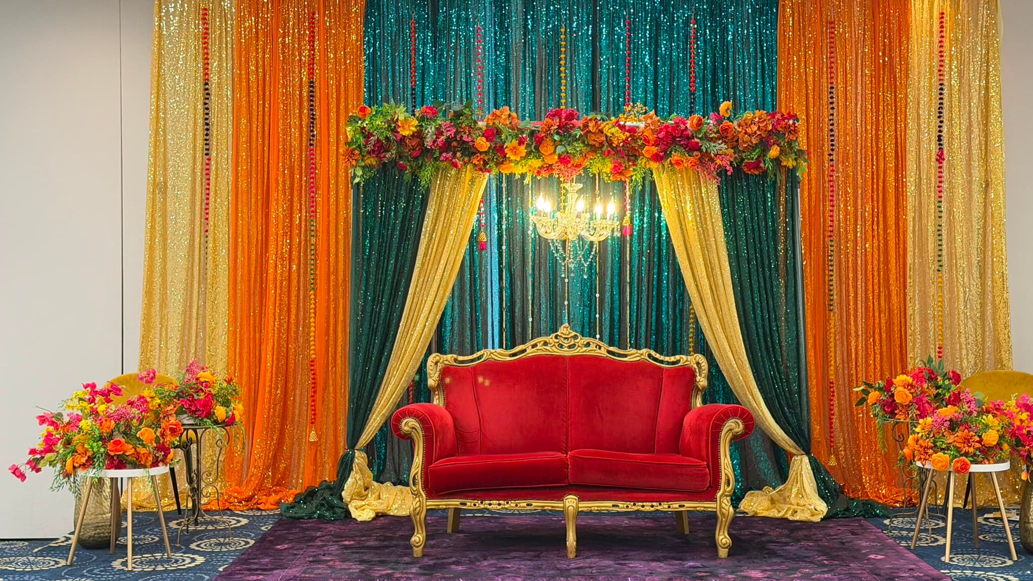 The wedding stage in the Bedfordshire Suite: gold, marigold and emerald sequin drapes behind a gilt-framed crimson velvet loveseat, under a chandelier hung with a garland of orange and pink flowers.